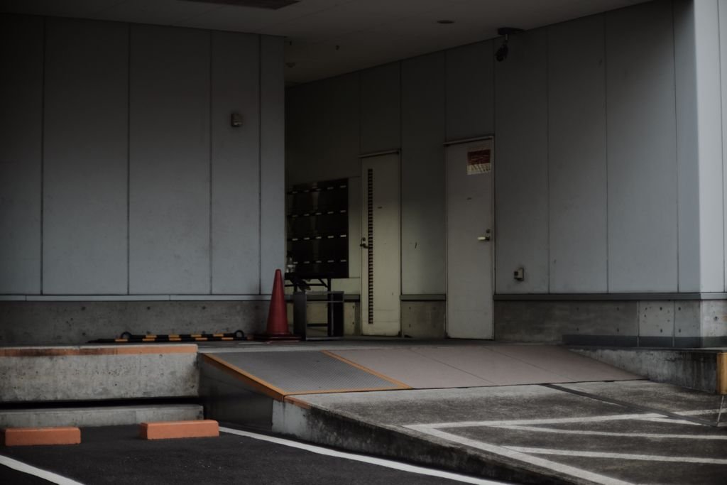 Muted industrial building entrance with a concrete driveway and orange traffic cones.